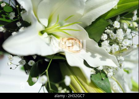 Wedding rings laying on bridal bouquet with white lily. Bridal accessories close up. Classic golden wedding bands. Stock Photo