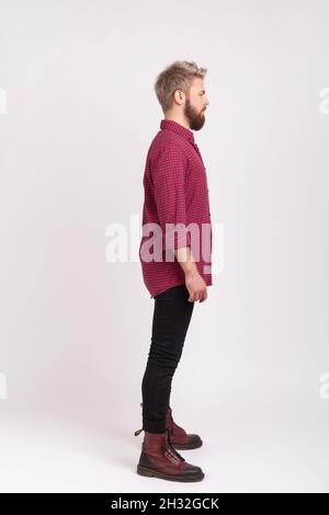 Side view full body length of serious confident bearded male wearing red plaid shirt and casual black pants posing, practicing modeling. Indoor studio shot isolated on gray background Stock Photo