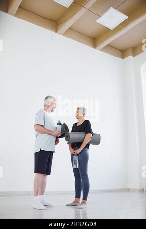 Senior couple with mats and water discussing yoga training together while standing in studio Stock Photo