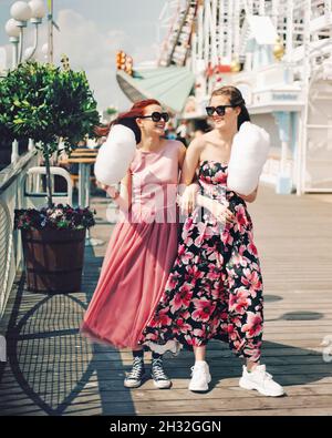 Happy girls with sunglasses and prom dresses holding cotton candy at amusement park. Teenagers in pink evening dresses on vacation. Birthday party ide Stock Photo