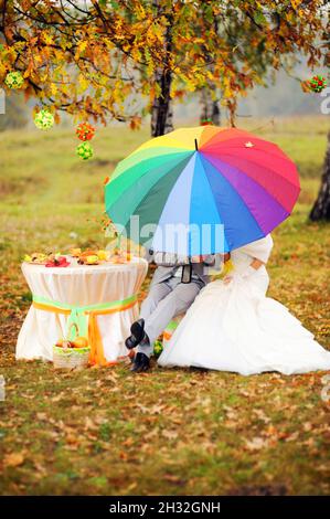 Autumn wedding. Outdoor portrait of newlyweds sitting under bright colorful umbrella (rainbow) in autumn park with yellow leaves. Stock Photo