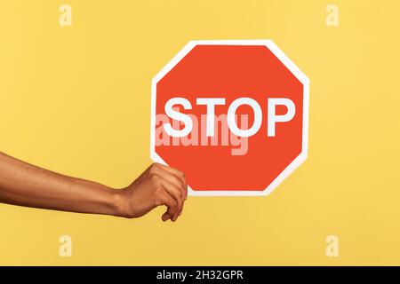 Closeup of male hand holding octagonal red Stop symbol, ban concept, arm showing traffic sign, prohibition concept, forbidden way, no access. Indoor studio shot isolated on yellow background. Stock Photo