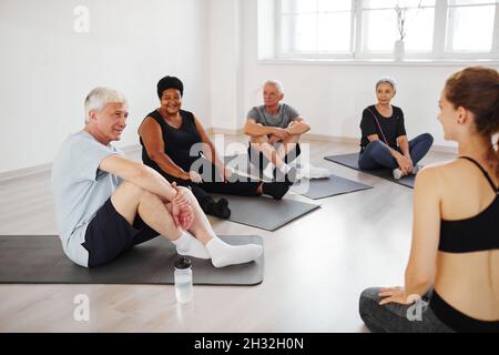 Senior people sitting on mats on the floor and practicing yoga together with young coach , they sitting and listening to her in class Stock Photo