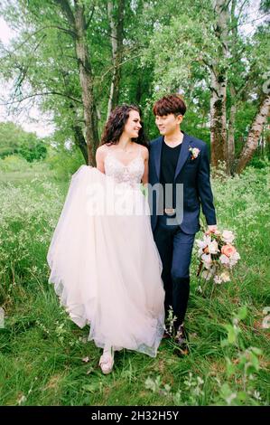 Smiling newlyweds walking and having fun in fresh green forest. Portrait of bride and groom. Outdoor spring wedding ceremony. International married co Stock Photo