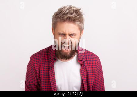 Portrait of displeased young man with beard screaming looking at camera, expressing anger and irritation, bad mood. Indoor studio shot isolated on gray background Stock Photo