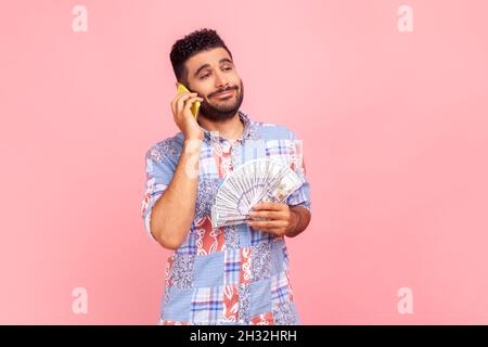 Portrait of attractive young adult man with beard in blue shirt standing, holding fan of money and talking on mobile phone, boasting with profit. Indoor studio shot isolated on pink background. Stock Photo