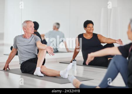 Senior man sitting on mat and doing relaxation exercises during yoga class with other people in studio Stock Photo