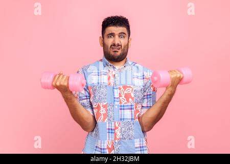Assertive funny man wearing casual style blue shirt rising hands holding dumbbells, pumping up muscles, having funny facial expression. Indoor studio shot isolated on pink background. Stock Photo