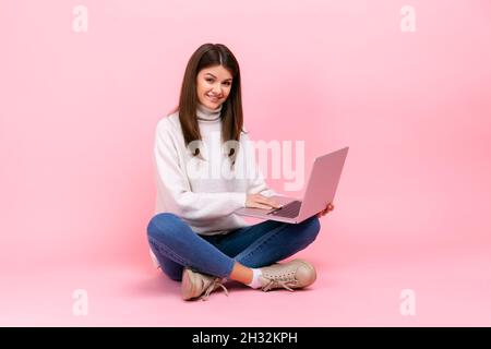 Positive satisfied female sitting with crossed legs on floor, holding laptop, looking at camera, wearing white casual style sweater. Indoor studio shot isolated on pink background. Stock Photo
