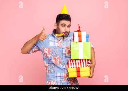 Portrait of bearded man with festive mood wearing party cone and casual shirt, showing thumb up to camera, holding stack of presents, and blowing horn. Indoor studio shot isolated on pink background. Stock Photo