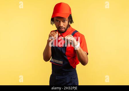 Portrait of aggressive workman with beard wearing uniform keeping fists clenched, ready for fight, profession of housekeeping maintenance. Indoor studio shot isolated on yellow background. Stock Photo
