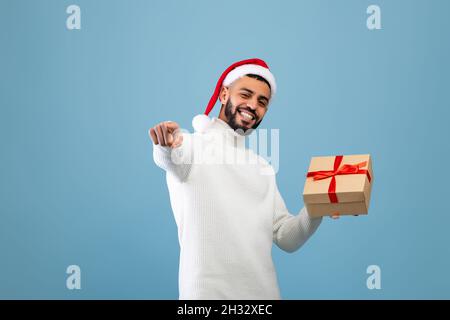 Joyful arab guy in Santa hat holding gift box and pointing finger at camera, standing over blue studio background. Happy man preparing Christmas or Ne Stock Photo