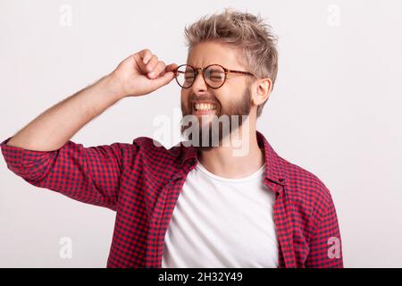 Portrait of funny extremely happy bearded man touching eyeglasses standing with closed eyes and laughing. Indoor studio shot isolated on gray background Stock Photo