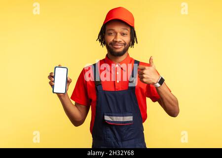 Satisfied handyman wearing overalls holding smart phone with blank screen for advertisement, showing thumb up, recommend service industry online app. Indoor studio shot isolated on yellow background. Stock Photo