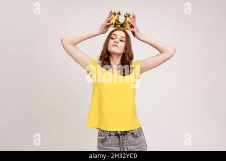 Portrait of selfish brown haired teenage girl in yellow T-shirt holding gold crown over head, having arrogance expression, privileged status. Indoor studio shot isolated on gray background. Stock Photo