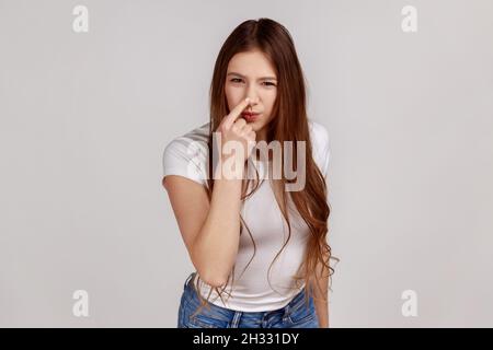 That's lie, you cheat. Disgruntled woman looking angrily at camera and showing lie gesture, touching nose meaning falsehood, wearing white T-shirt. Indoor studio shot isolated on gray background. Stock Photo