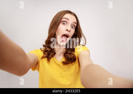 Portrait of excited surprised teenager girl wearing yellow casual T shirt taking selfie, looking at camera with open mouth POV, point of view of photo. Indoor studio shot isolated on gray background. Stock Photo