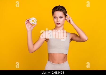 Photo of unsure doubtful lady hold doughnut decide diet wear grey top isolated yellow color background Stock Photo