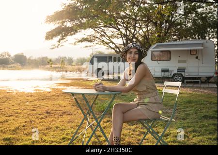 Cheerful young asian woman sitting and smiling in campsite on lakeside and camper van parked at the evening Stock Photo