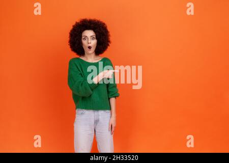 Shocked surprised woman with Afro hairstyle wearing green casual style sweater pointing away with finger, keeps mouth widely open, astonishing ad. Indoor studio shot isolated on orange background. Stock Photo