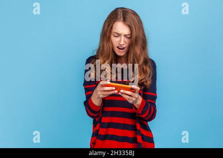 Portrait of woman wearing striped casual style sweater, holding smart phone in hands and playing video games with concentrated look, loosing level. Indoor studio shot isolated on blue background. Stock Photo