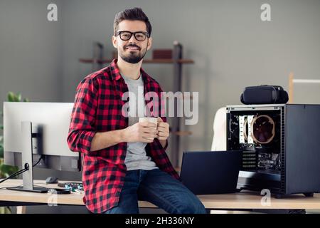 Portrait of nice attractive smart clever cheerful guy sitting on table drinking beverage at work place station indoors Stock Photo