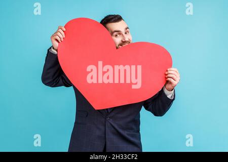 Positive man wearing official style suit hiding behind big red heart and looking at camera with playful eyes, holding symbol of love and affection. Indoor studio shot isolated on blue background. Stock Photo