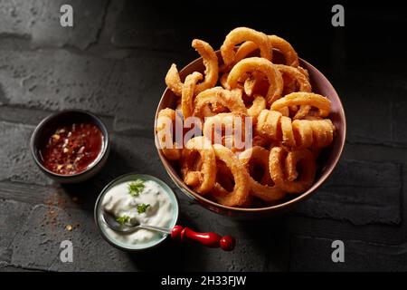 Top view of a bowl of onion rings and curled potatoes placed on a black table near two tasty sauces Stock Photo