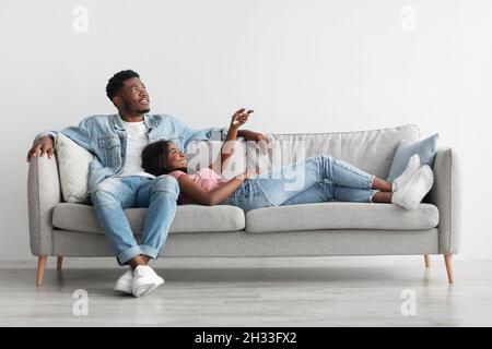 Cheerful Black Lady Holding Remote Controller, Opening Air Conditioner At Home While Relaxing On Comfortable Couch In Living Room, Happy Woman Adjusti Stock Photo