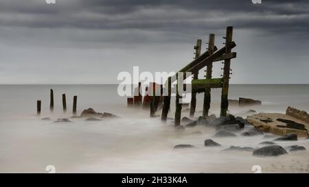 The reamins of the sea defences below the rapidly eroding cliffs of Happisburgh (Haisbro) on the North Norfolk coast rise like sentinels from the sea. Stock Photo