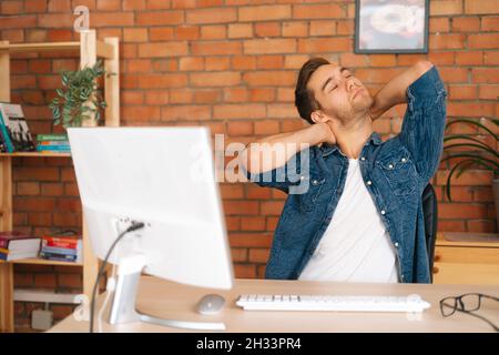 Portrait of exhausted young business man experiences severe pain in neck, rubbing it to relieve muscle tension, sitting at desk. Stock Photo