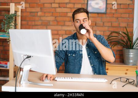 Front view of busy young freelance programmer male drinking coffee from cup working on desktop computer, sitting at desk in home office. Stock Photo