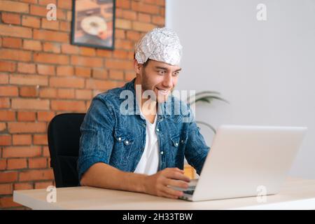 Medium shot of crazy male conspiracy theorist wearing aluminum foil protect brain watching shocked online video content using laptop computer. Stock Photo