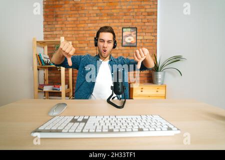 Front view of negative angry male blogger streaming live podcast using professional microphone showing double thumbs down Stock Photo