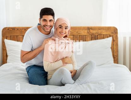 Caring arab husband massaging his pregnant wife's shoulders at home, sitting together on bed and smiling at camera, free space. Man giving massage to Stock Photo