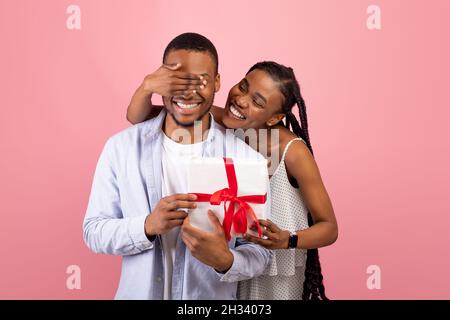 Happy Birthday Or Anniversary. Smiling black woman covering her boyfriend's eyes, greeting and giving him wrapped gift box, standing isolated on pink Stock Photo