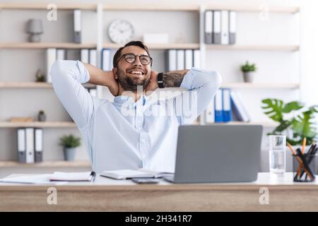 Satisfied middle aged caucasian guy with beard in glasses resting from work at laptop in office or living room interior. Successful business and deal Stock Photo