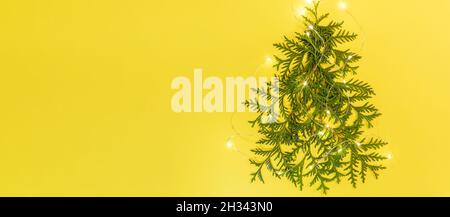 Christmas holiday banner. Xmas tree made from green arborvitae branches with luminous Xmas lights on yellow background. Alternative Xmas Tree. Stock Photo