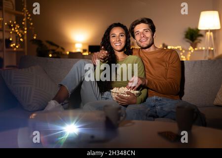 Happy Couple Relaxing Watching Movie Using Projector Eating Popcorn And Embracing Sitting Together On Sofa In Living Room At Home. Boyfriend And Girlf Stock Photo