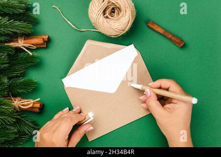 top view, process of writing letter to Santa Claus on craft envelope on green table with decorations of Christmas trees, cinnamon and jute rope Stock Photo