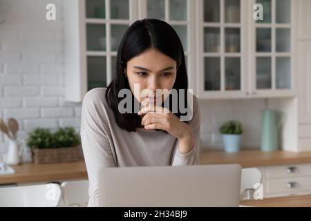 Serious focused Asian student using laptop in kitchen Stock Photo