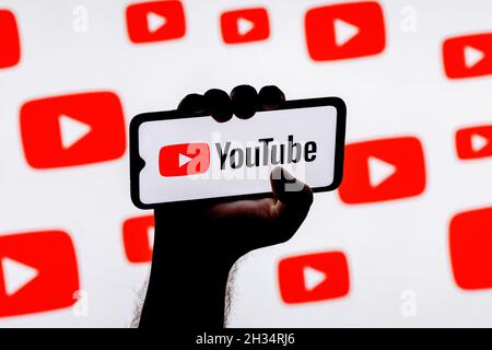Smartphone with the Youtube social media platform logo on the screen in a clenched hand on the background of Youtube logos Stock Photo