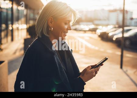 Portrait of a Caucasian woman smiling while texting friends. Blond hair in the setting sun. Young woman receive some nice news on her cellphone and Stock Photo