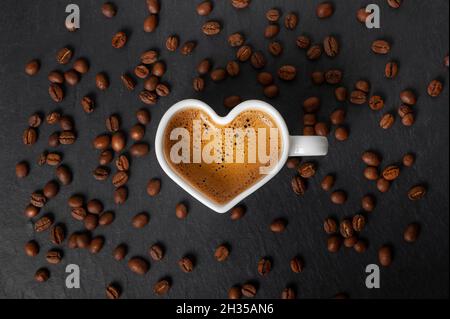 white cup in the shape of a heart with coffee and beautiful foam on a black stone background with scattered coffee beans. view from above. copy space Stock Photo