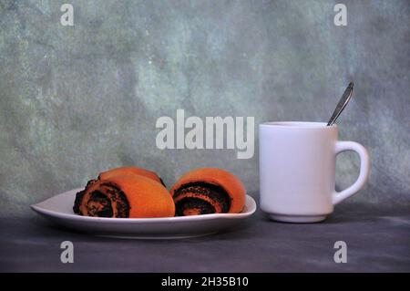 Fresh rolled poppy seed buns on a white plate and a cup of hot black tea on a gray background. Close-up. Stock Photo