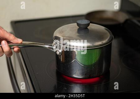 Hand holds handle of stainless steel pot on induction stove Stock Photo