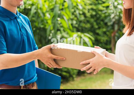 Asian young delivery man courier in uniform hold parcel post boxes service shipment woman customer receiving accepting parcel from delivery at front h Stock Photo