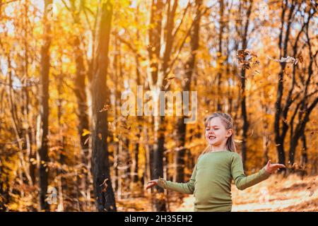 Small happy smiling little caucasian child girl playing with yellow leaves in autumn forest or garden around trees. Kid throws leaves up and they are Stock Photo