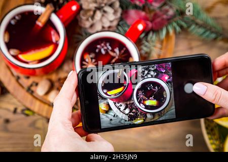 Female hands holding smartphone and taking photo of two colorful cups of traditional Christmas drink mulled red spiced wine with cinnamon stick, apple Stock Photo