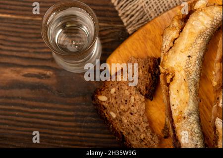 A small fried fish on a wooden board, pieces of bread and a glass of vodka, close-up, selective focus Stock Photo